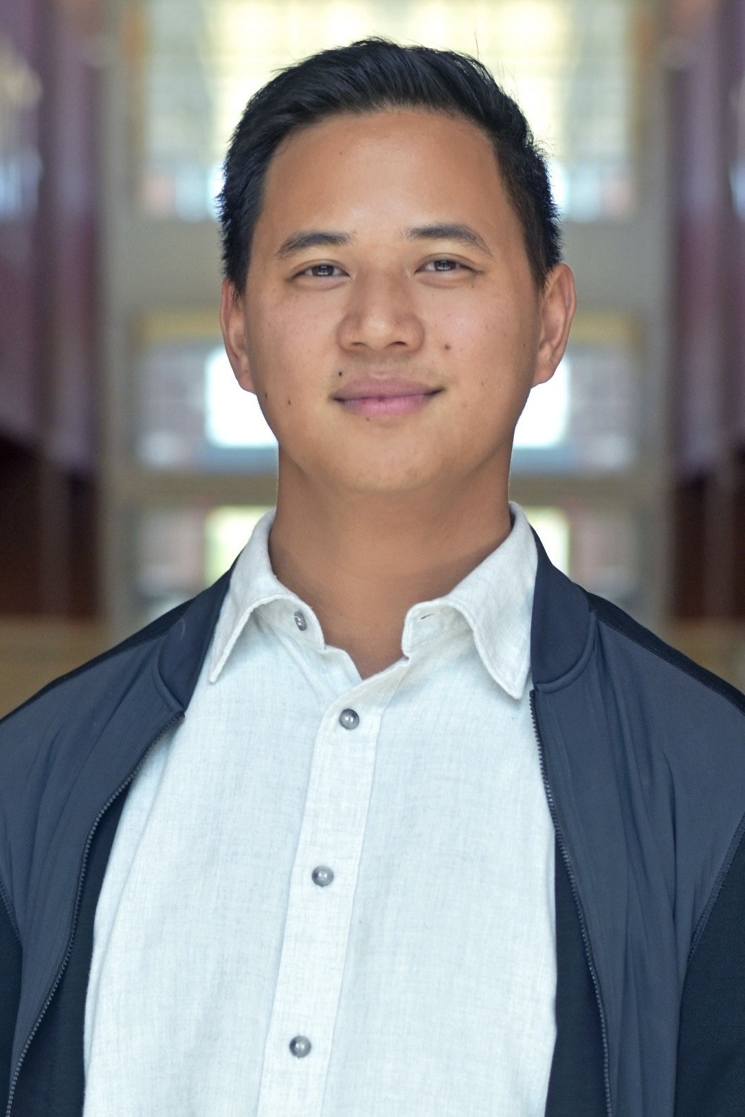 Headshot person with short black hair in a white button-down under an blue zip-up jacket.