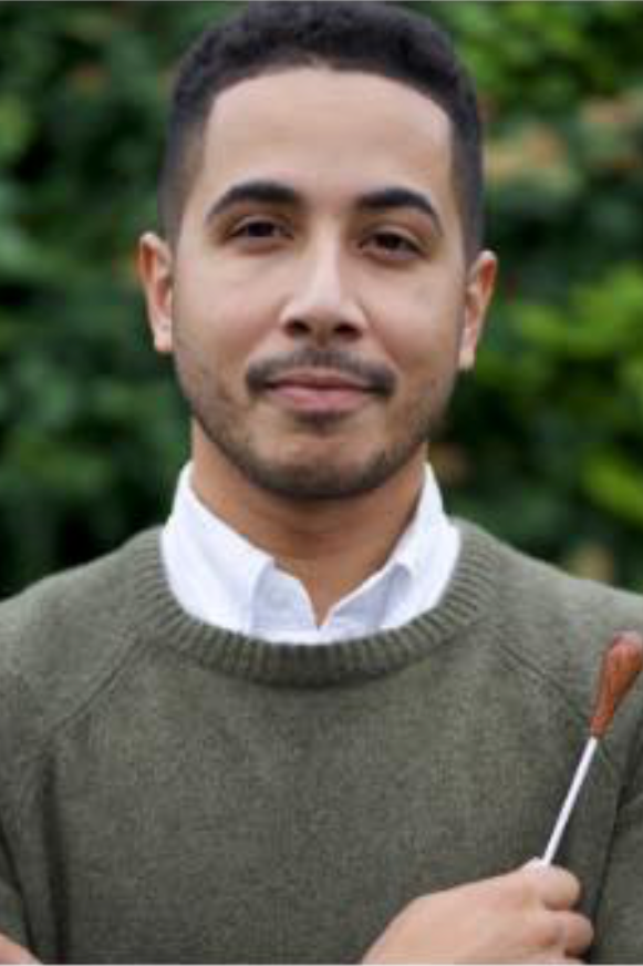 Headshot of hispanic person with short black hair in a white button-down under an olive sweater. He holds a baton and stands in front of a background of green trees