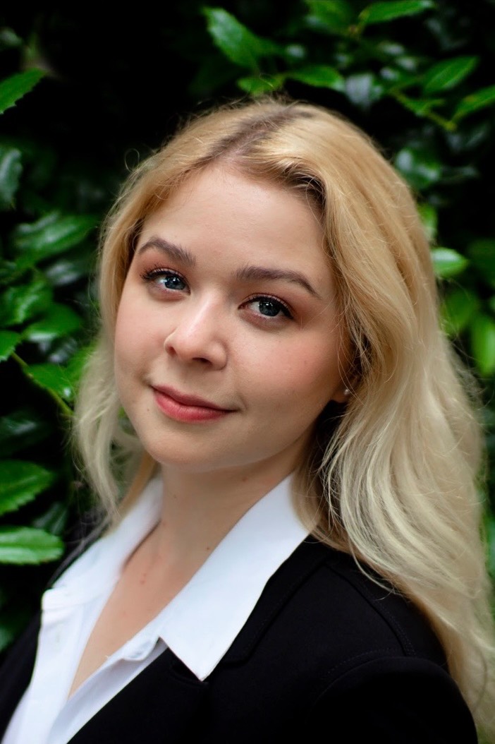 Headshot of a white person with long blonde hair in a white button-down under a black blazer with nature background