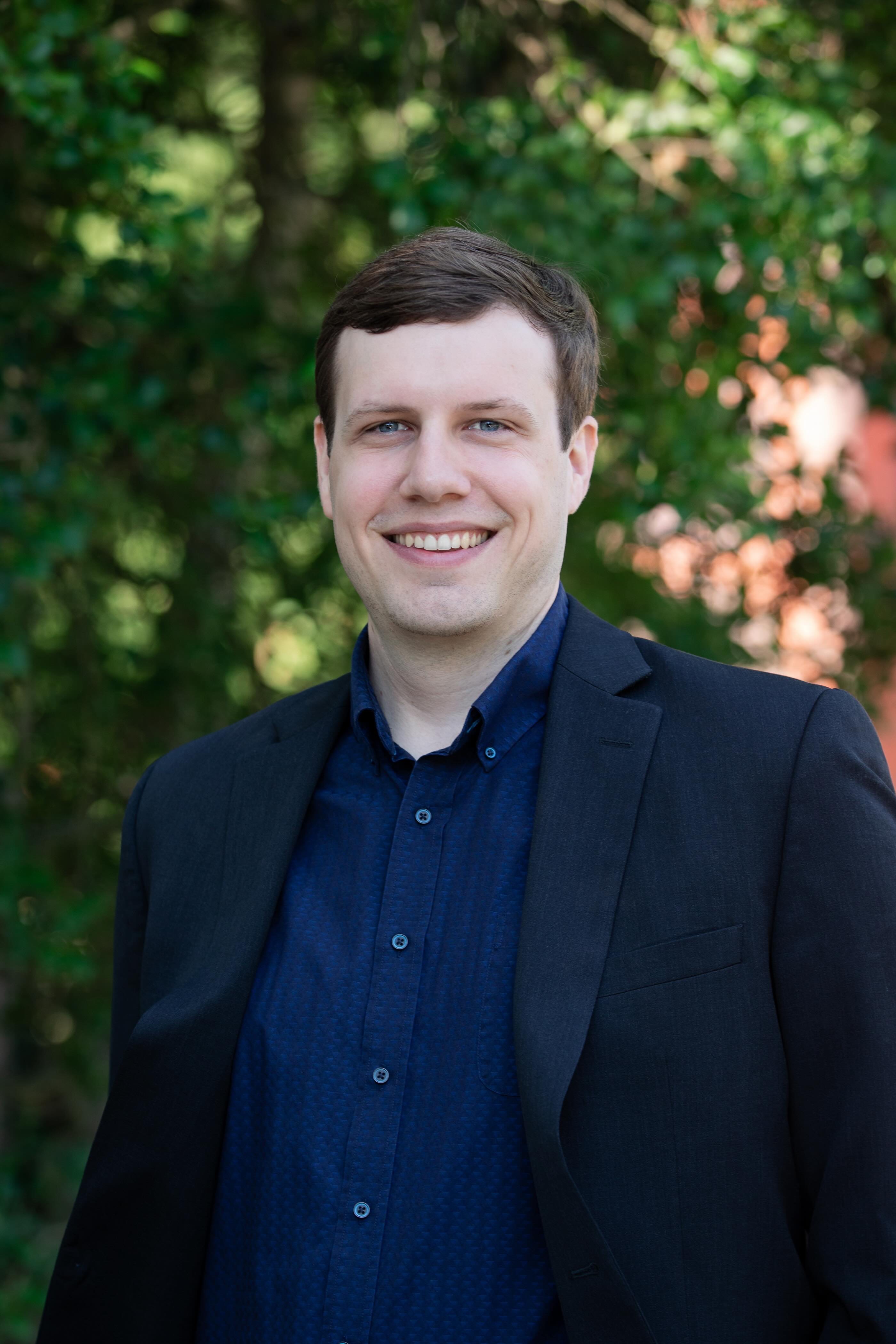 Headshot of a white person with short brown hair in a blue patterned button-down under a navy blazer and a tree in the background