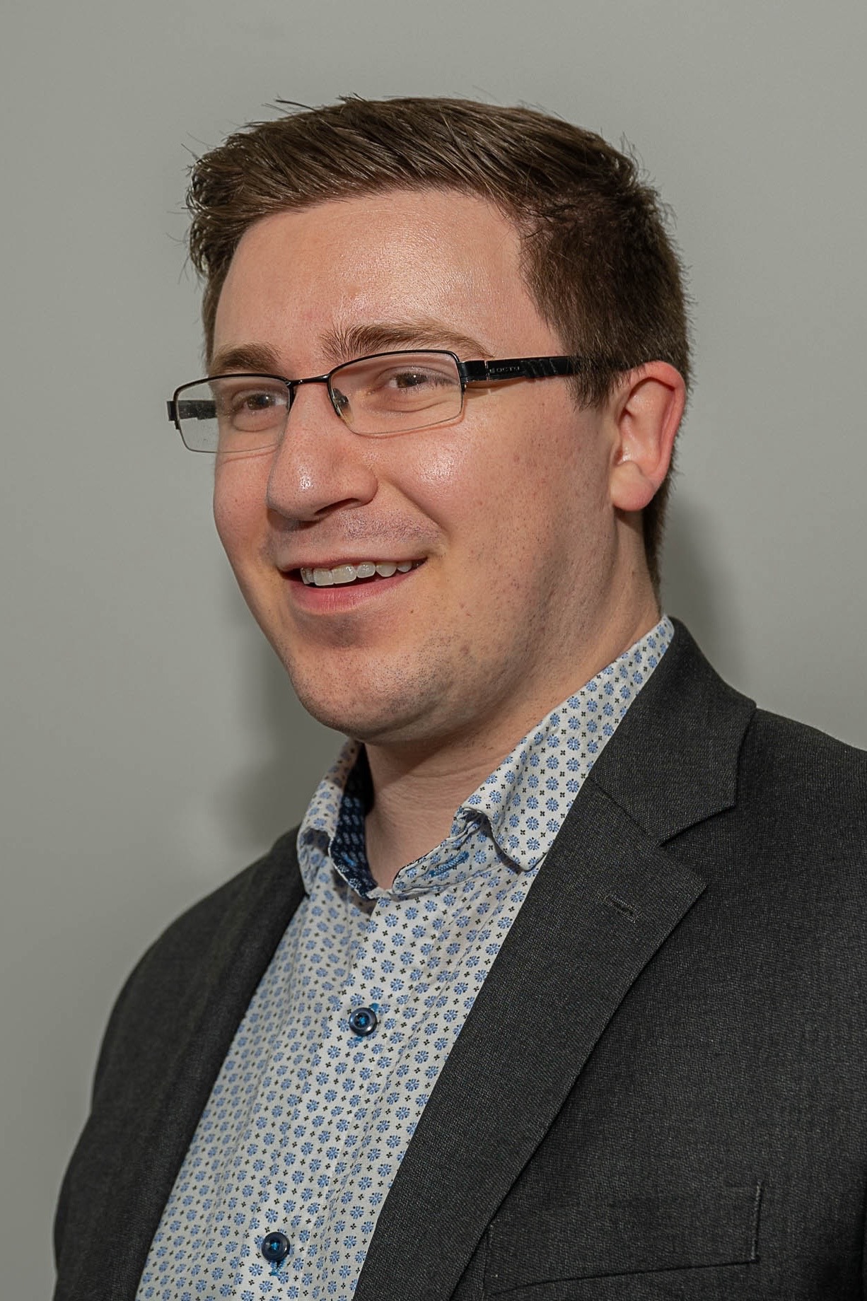 Headshot of white person with short brown hair and glasses, wearing a blue-on-white patterned shirt under a charcoal blazer