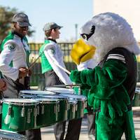 Scrappy plays the tenor drums while a drumline student watches with amusement