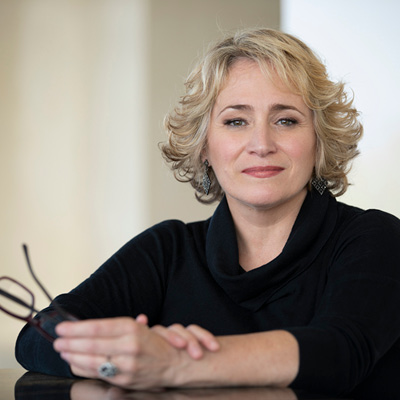 Image is Ms. Racette seated with her arms folded on a table holding a pair of eyeglasses wearing a black outfit.