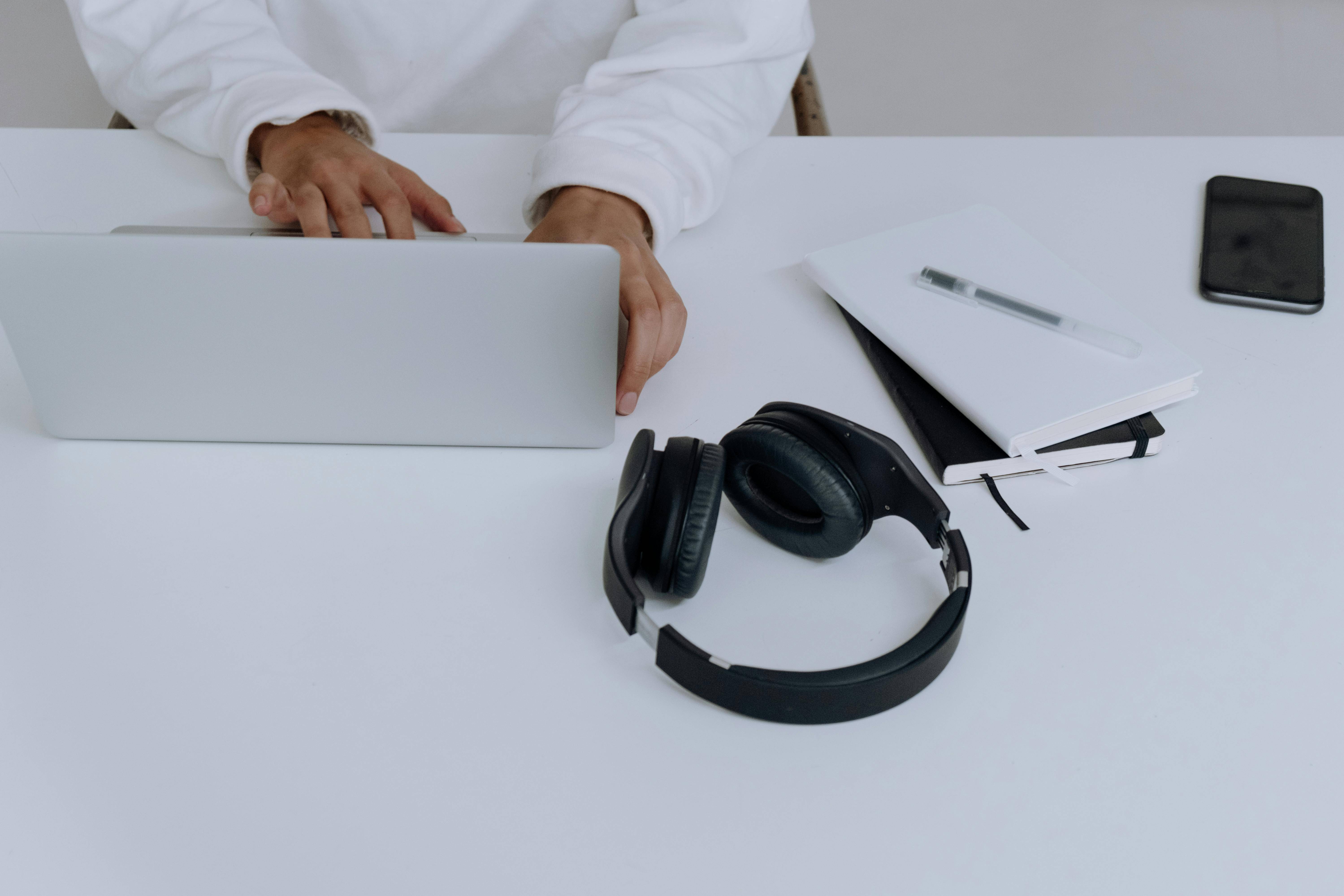 Person at desk with laptop, headphones, a journal, a notepad, and phone.