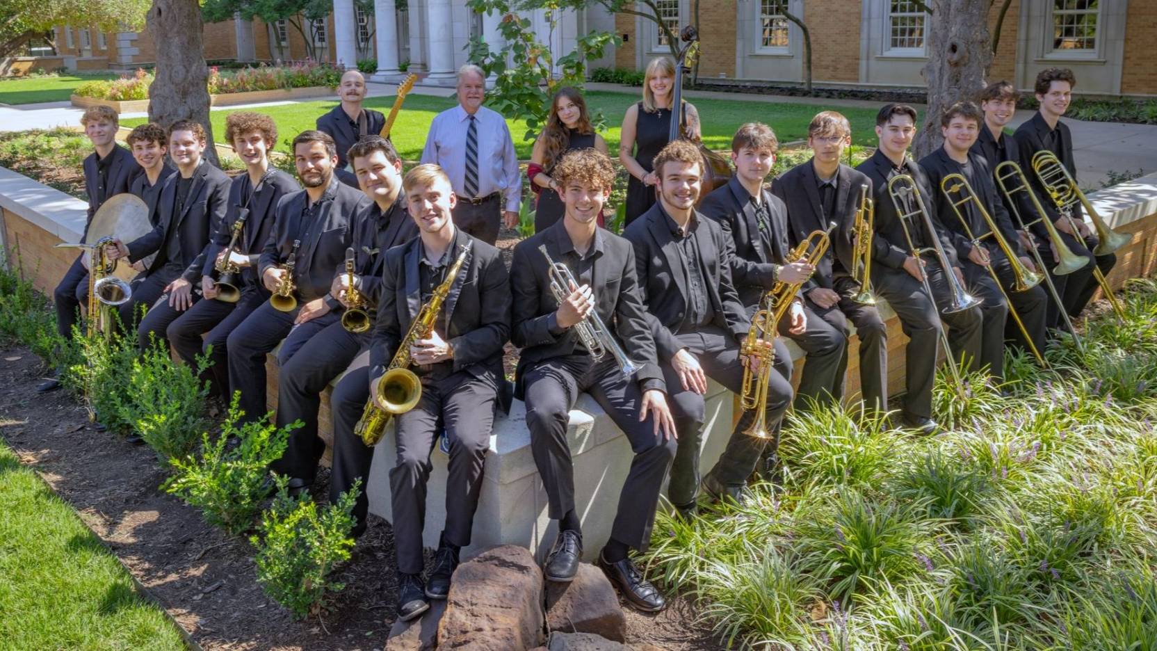 The Two O'Clock Lab Band sits on a short wall in dark suits holding their instruments.