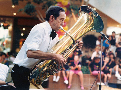 A photo of Everett playing tuba in front of a group of students.  He is wearing a bowtie and white shirt.